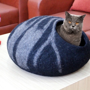 May include: A gray cat sits inside a blue and gray felt cat bed. The bed is round and has a hole in the side for the cat to enter. The cat is looking directly at the camera.