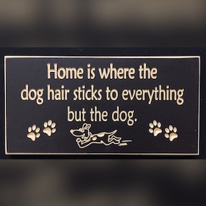 May include: Black wooden sign with the text "Home is where the dog hair sticks to everything but the dog." in gold lettering. The sign also features two gold paw prints on each side and a gold cartoon dog running in the centre.