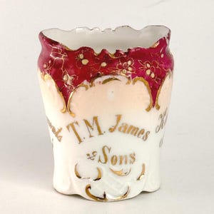 May include: A small, antique white porcelain cup with a flared rim. The rim is decorated with a red floral pattern and gold accents. The cup has the text "T.M. James & Sons" in gold lettering.