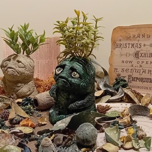 May include: Two small green and gray ceramic planters with sad faces and green plants growing out of the top. The planters are sitting on a wooden surface with dried flowers and leaves scattered around them. A vintage advertisement for a Christmas exhibit is visible in the background.
