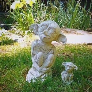 Puede incluir: Dos estatuas de jardín de hormigón gris de una criatura sentada con forma de duende y orejas grandes. Una estatua es más grande que la otra. Las estatuas están en una zona de césped con follaje verde y flores amarillas en el fondo.