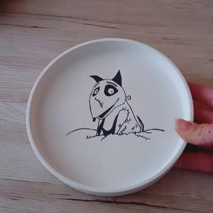 Könnte beinhalten: Ein runder, cremefarbener Keramikteller mit einer schwarz-weißen Illustration eines Cartoon-Hundes mit genähten Merkmalen. Der Hund sitzt auf einem kleinen Hügel. Der Teller hat einen leicht erhöhten Rand.