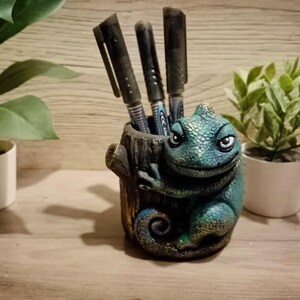 Könnte beinhalten: Ein skurriler Stifthalter in Form eines blau-grünen Chamäleons, das einen Baumstamm umklammert. Drei schwarze Stifte befinden sich darin. Der Halter steht auf einer Holzoberfläche, flankiert von zwei kleinen Topfpflanzen.