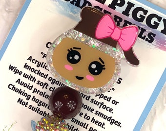 May include: A blue piggy badge reel with a sparkly brown pig face, pink bow, and brown hair. The reel has a clear plastic string and a metal clip. The card reads "Blue Piggy Badge Reels" and includes instructions for care and use.