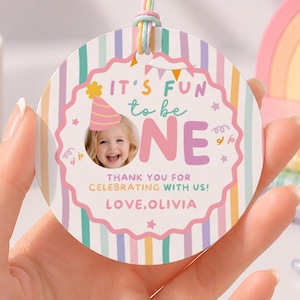 Puede incluir: Una etiqueta de regalo redonda de papel con fondo de rayas pastel, una foto de un niño con gorro de fiesta, el texto IT'S FUN to be ONE, THANK YOU FOR CELEBRATING WITH US! y LOVE, OLIVIA, sujeta a un cordel multicolor.