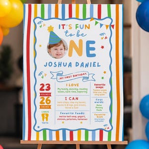 May include: A first birthday milestone sign on a wooden easel featuring a photo of a child in a party hat, the text IT'S FUN to be ONE, JOSHUA DANIEL, MY FIRST BIRTHDAY, 23 POUNDS, 26 INCHES, 5 TEETH, and lists of favorite things.