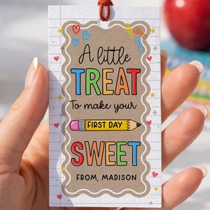 May include: A rectangular paper gift tag with a scalloped border on a lined background. The text reads A little TREAT To make your FIRST DAY SWEET FROM, MADISON. It features a pencil graphic, colorful hearts, and a red string at the top.