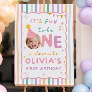 Pode incluir: Uma placa retangular de boas-vindas de aniversário num cavalete de madeira com a foto de uma criança com chapéu de festa, riscas verticais pastel e o texto: IT'S FUN to be ONE welcome to OLIVIA'S FIRST BIRTHDAY, rodeada por um arco de balões.