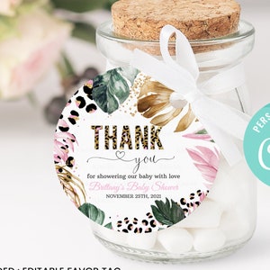 Puede incluir: Una etiqueta de favor redonda de 5 cm x 5 cm con un tema tropical. La etiqueta presenta un fondo de purpurina dorada con hojas verdes, flores rosas y detalles de estampado de leopardo. El texto en la etiqueta dice "Gracias por consentir a nuestro bebé con amor, Baby Shower de Brittany, 25 de noviembre de 2021".