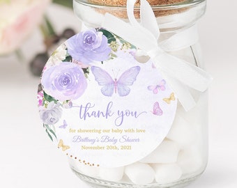 May include: A small glass jar filled with white candy, tied with a white ribbon and a round thank you tag. The tag features a watercolor design of purple flowers and butterflies with the text "thank you for showering our baby with love Brittany's Baby Shower November 20th, 2021".