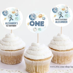 Op de afbeelding: Drie cupcakes versierd met ronde, witte, blauwe en gouden cupcake-toppers met een ruimtethema. De toppers tonen een astronaut, planeten en de woorden "Happy Birthday Alexander", "One" en "Alexander's 1st Birthday".