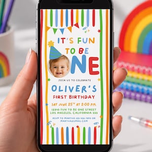 May include: A smartphone displaying a digital first birthday invitation with a colorful striped border, a photo of a child wearing a party hat, and the text: It's Fun To Be One, Oliver's First Birthday, Sat June 25th at 2:00 PM, 12345 Fun To Be One Street.