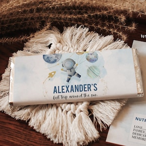Puede incluir: Un envoltorio de barra de chocolate personalizado con una ilustración de astronauta azul y blanco y el texto "ALEXANDER'S first trip around the sun".