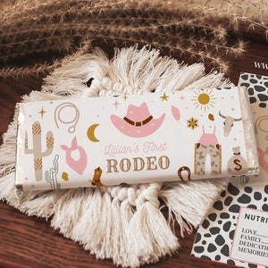May include: A white chocolate bar wrapper with a pink cowboy hat, cactus, and stars. The text "Lillian's First Rodeo" is printed on the wrapper. The wrapper is on a brown wooden surface.