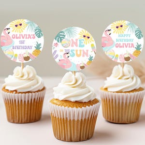 May include: Three cupcakes with white frosting, each topped with a circular decoration. The decorations feature a flamingo, sun, and tropical elements. Text on the toppers reads "Olivia's 1st Birthday", "One in the Sun", and "Happy Birthday Olivia".