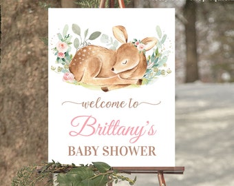 May include: A white welcome sign with a watercolor illustration of a fawn sleeping in a bed of pink flowers and green leaves. The text "welcome to Brittany's Baby Shower" is written in pink and black cursive font.