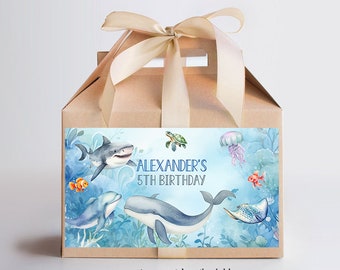 Op de afbeelding: Een bruine traktatiebox met een blauw en wit label met een aquarel-illustratie van zeedieren, waaronder een haai, een dolfijn, een walvis, een schildpad, een kwal en een vis. Het label bevat ook de tekst "Alexander's 5th Birthday".