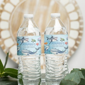 May include: Two clear plastic water bottles with blue and white labels. The labels feature a watercolor illustration of a shark, turtle, fish, and whale. The text on the label reads "Alexander's 5th Birthday".