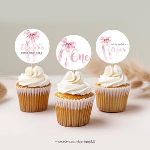 May include: Three cupcakes with pink and white frosting topped with round paper cupcake toppers. The toppers feature a pink watercolor bow and the text "Elizabeth's First Birthday", "One", and "Happy Birthday Elizabeth".