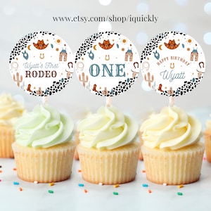 May include: Three round cupcake toppers with a cowboy theme. The toppers feature a white background with brown and black spots, a blue star, a yellow sun, a cactus, a horseshoe, a cowboy hat, and a pair of cowboy boots. The text on the toppers reads "Wyatt's First Rodeo", "One", and "Happy Birthday Wyatt".