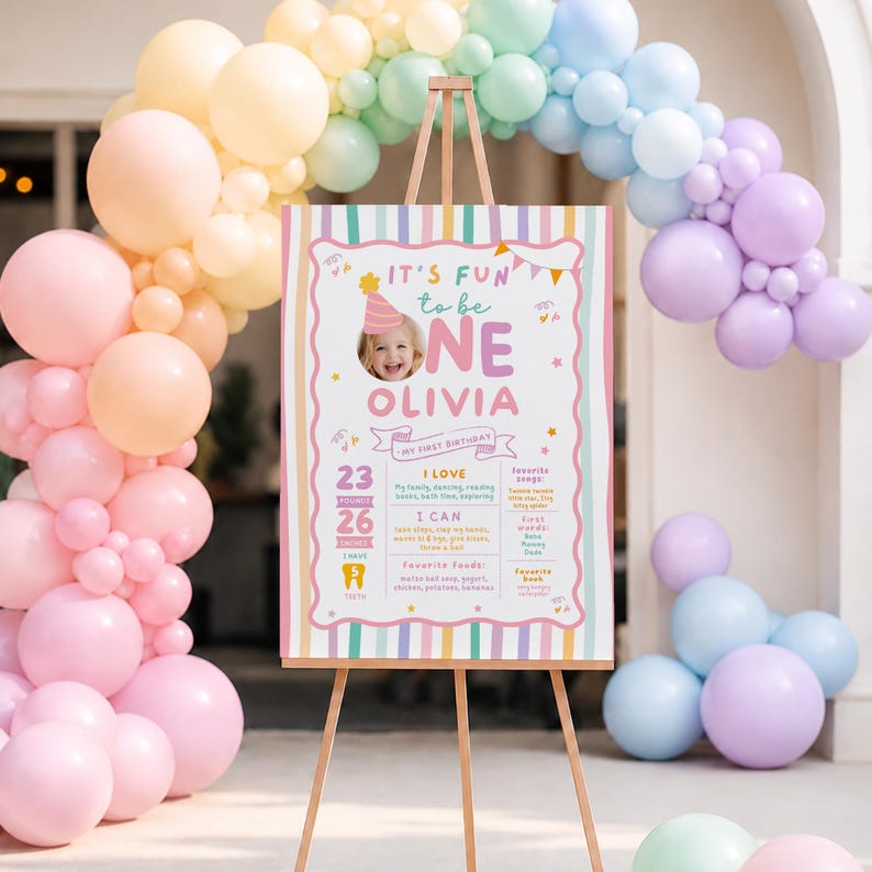 Pu&ograve; includere: Un cartello di compleanno rettangolare su un cavalletto in legno, con strisce verticali color pastello, una foto di un bambino con cappellino da festa e testo che include 'IT'S FUN to be ONE OLIVIA', '10,4 kg', '66 cm' e '5 DENTI'.