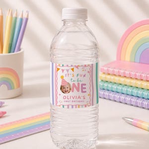 Pu&ograve; includere: Una bottiglia d'acqua in plastica trasparente con un'etichetta personalizzata che mostra la foto di un bambino con un cappellino da festa, strisce verticali colorate e il testo IT'S FUN to be ONE OLIVIA'S FIRST BIRTHDAY con stelline.