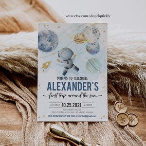 May include: A blue and white invitation with gold accents for a child's first trip around the sun party. The invitation features an astronaut holding balloons shaped like planets. The text on the invitation reads "Join us to celebrate Alexander's first trip around the sun. Saturday 10.25.2021 2:00PM. Alexander's House 2345 Floral Blush Ave Las Vegas Nevada. Rsvp to Martha 567-890-1234 or martha@gmail.com"