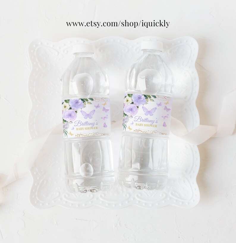 May include: Two clear plastic water bottles with purple and gold labels. The labels feature a floral design with butterflies and the text "Brittany's Baby Shower".