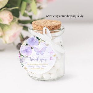 May include: Purple and white floral thank you tag with butterflies and gold glitter. The tag is attached to a small glass jar filled with white candy. The text on the tag reads "thank you for showering our baby with love Brittany's Baby Shower November 20th, 2021".