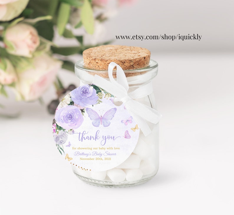 May include: Purple and white floral thank you tag with butterflies and gold glitter. The tag is attached to a small glass jar filled with white candy. The text on the tag reads "thank you for showering our baby with love Brittany's Baby Shower November 20th, 2021".
