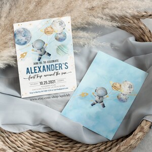 May include: A blue and white invitation card with a watercolor illustration of an astronaut holding balloons shaped like planets. The text on the card reads "Join us to celebrate Alexander's first trip around the sun" and includes the date and time of the event.