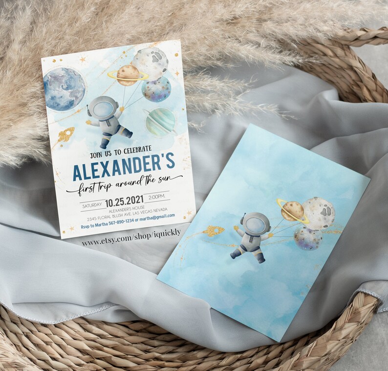 May include: A blue and white invitation card with a watercolor illustration of an astronaut holding balloons shaped like planets. The text on the card reads "Join us to celebrate Alexander's first trip around the sun" and includes the date and time of the event.