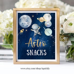 May include: A gold-framed sign with a blue background and a cartoon astronaut holding planets. The text "Astro Snacks" is written in gold lettering.