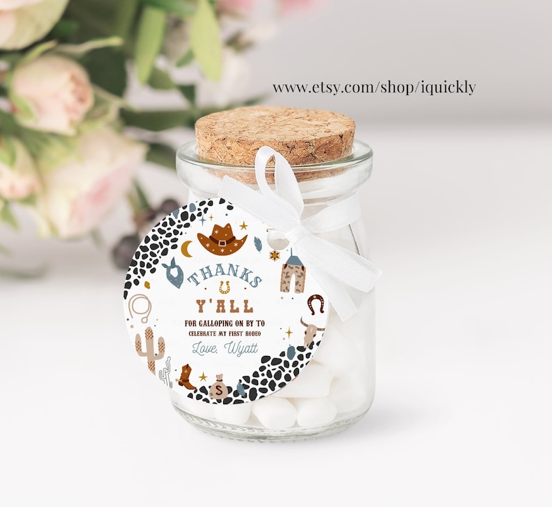 May include: Round favor tag with a cowboy theme. The tag is white with black and brown spots. It features a cowboy hat, a horseshoe, a cactus, and the text "Thanks Y'all" and "For Galloping On By To Celebrate My First Rodeo".