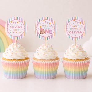 Pu&ograve; includere: Tre cupcake in pirottini di carta a righe arcobaleno, guarniti con glassa bianca e zuccherini colorati. Ogni cupcake presenta un topper rotondo di carta su uno stecchino di legno. I topper mostrano righe pastello, festoni e il testo Olivia's First Birthday, It's Fun to be One e Happy Birthday Olivia.