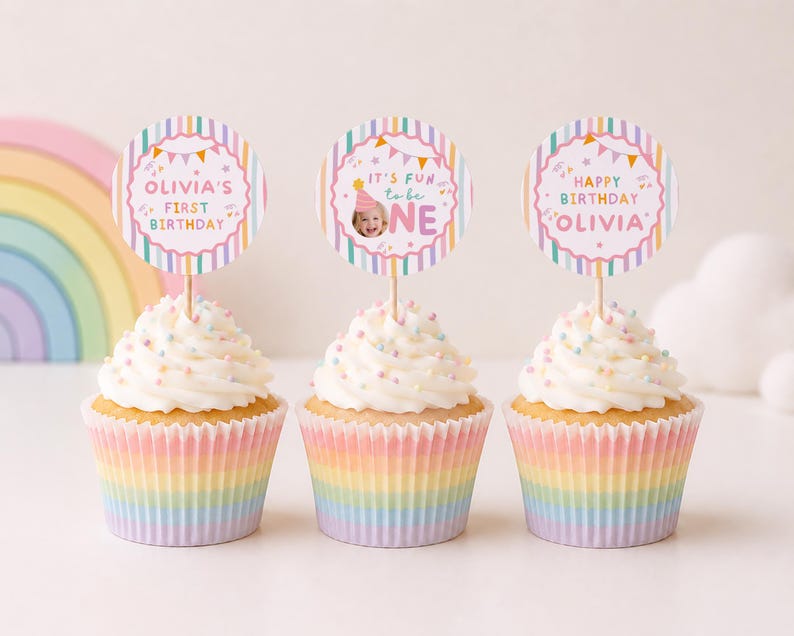 Pu&ograve; includere: Tre cupcake in pirottini di carta a righe arcobaleno, guarniti con glassa bianca e zuccherini colorati. Ogni cupcake presenta un topper rotondo di carta su uno stecchino di legno. I topper mostrano righe pastello, festoni e il testo Olivia's First Birthday, It's Fun to be One e Happy Birthday Olivia.