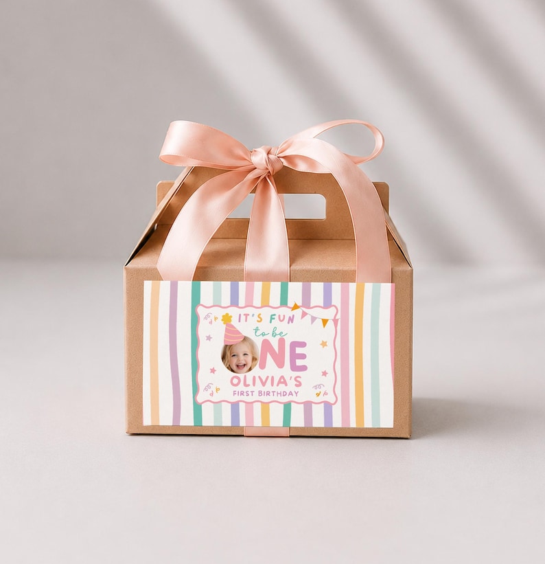 Pu&ograve; includere: Una scatola di cartone marrone con manico, legata con un nastro di raso rosa chiaro. La parte anteriore presenta un'etichetta con strisce verticali color pastello e una grafica centrale con la foto di un bambino, il testo "IT'S FUN to be ONE" e "OLIVIA'S FIRST BIRTHDAY".