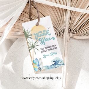May include: A white tag with a beach-themed design. The tag features a yellow sun, palm trees, and surfboards. Text on the tag reads "Thank You" and "for riding the waves with me!" with the name "Riley" at the bottom.