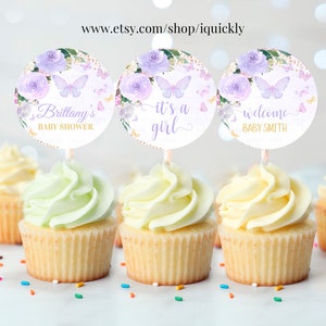 May include: Three cupcake toppers with purple flowers, butterflies, and gold glitter. The toppers say "Brittany's Baby Shower", "It's a Girl", and "Welcome Baby Smith".