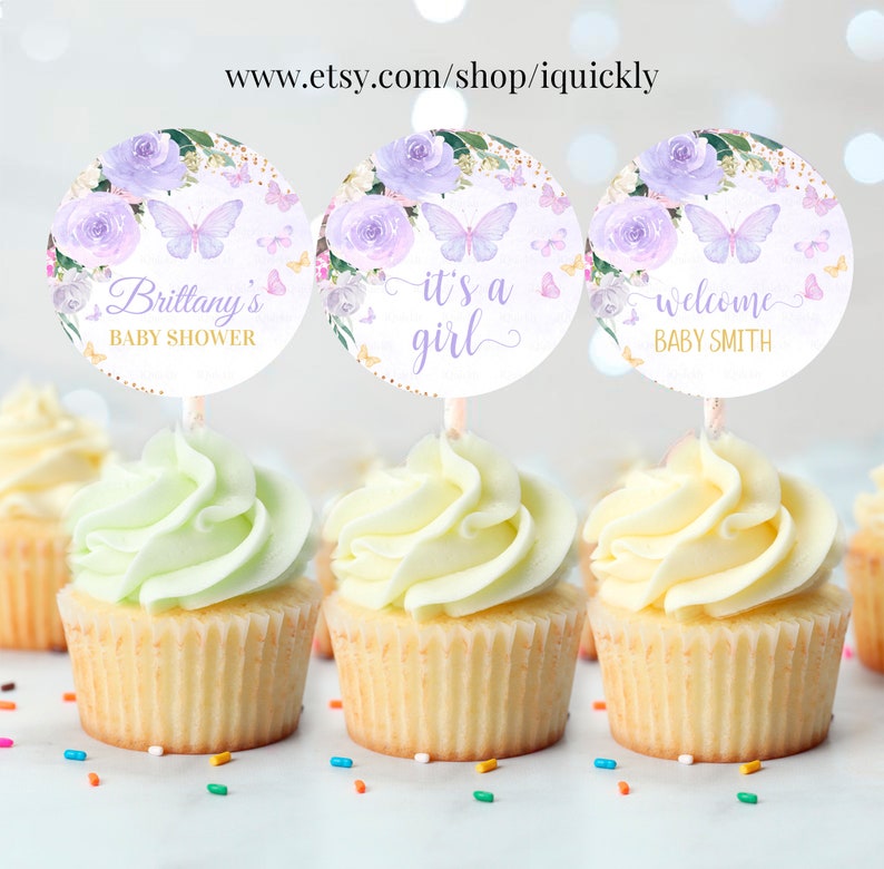 May include: Three cupcake toppers with purple flowers, butterflies, and gold glitter. The toppers say "Brittany's Baby Shower", "It's a Girl", and "Welcome Baby Smith".
