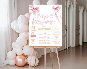 May include: A pink and gold first birthday milestone sign featuring a watercolour bow design. The sign includes the name "Elizabeth" and details about her first year, including age, height, weight, and favourite foods.