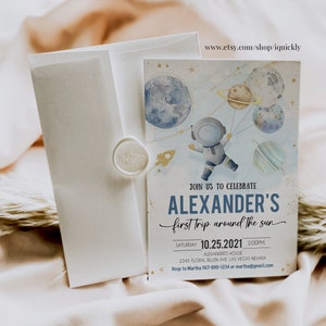 May include: A blue and white invitation card with a watercolor illustration of an astronaut holding balloons shaped like planets. The text on the card reads "Join us to celebrate Alexander's first trip around the sun" and includes the date and time of the event.