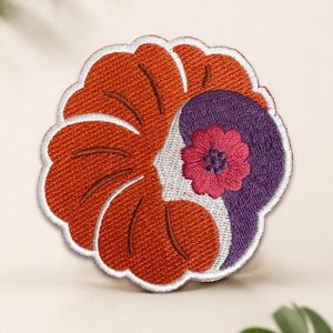 May include: Embroidered patch featuring a stylized floral design. The patch has a vibrant color scheme, with a large orange petal section and a purple section with a pink flower. The patch is outlined in white.