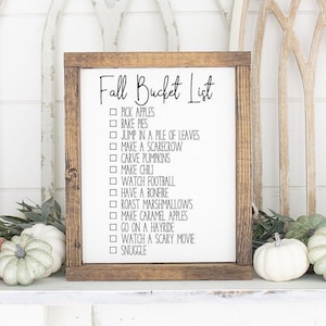 May include: A wooden sign with the text "Fall Bucket List" and a checklist of fall activities, including picking apples, baking pies, jumping in a pile of leaves, making a scarecrow, carving pumpkins, making chili, watching football, having a bonfire, roasting marshmallows, making caramel apples, going on a hayride, watching a scary movie, and snuggling.