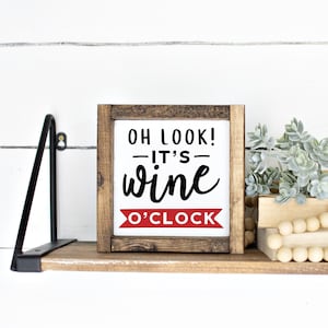 May include: A wooden sign with the text "Oh look! - It's - wine o'clock" in black lettering on a white background. The sign is framed in brown wood and has a red banner at the bottom with the text "o'clock". The sign is sitting on a wooden shelf with a small plant and a string of wooden beads.