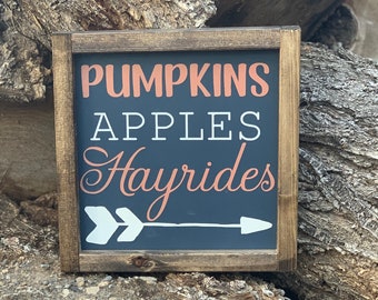 May include: A wooden sign with a black background and white text that reads "Pumpkins Apples Hayrides" with an orange arrow pointing to the right.