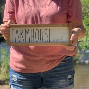 K&ouml;nnte beinhalten: Ein rechteckiges Holzschild mit den Worten "FARMHOUSE ish" in schwarzer Schrift. Das Schild hat einen wei&szlig;en Hintergrund und einen braunen Holzrahmen. Das Schild wird von einer Person gehalten, die ein rosa T-Shirt und Jeansshorts tr&auml;gt.