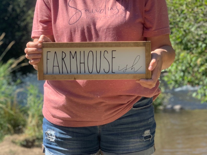 K&ouml;nnte beinhalten: Ein rechteckiges Holzschild mit den Worten "FARMHOUSE ish" in schwarzer Schrift. Das Schild hat einen wei&szlig;en Hintergrund und einen braunen Holzrahmen. Das Schild wird von einer Person gehalten, die ein rosa T-Shirt und Jeansshorts tr&auml;gt.