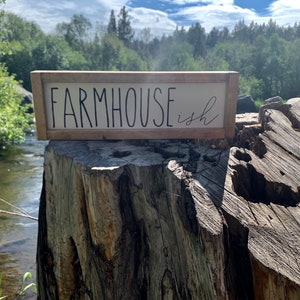 K&ouml;nnte beinhalten: Ein rechteckiges Holzschild mit den Worten "FARMHOUSE ish" in schwarzer Schrift. Das Schild hat einen hellen Hintergrund und einen braunen Rahmen. Das Schild steht auf einem Baumstumpf mit einem Fluss und B&auml;umen im Hintergrund.