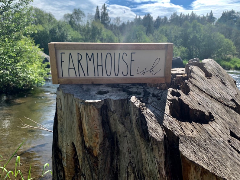 K&ouml;nnte beinhalten: Ein rechteckiges Holzschild mit den Worten "FARMHOUSE ish" in schwarzer Schrift. Das Schild hat einen hellen Hintergrund und einen braunen Rahmen. Das Schild steht auf einem Baumstumpf mit einem Fluss und B&auml;umen im Hintergrund.